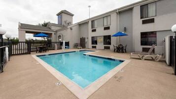 A rectangular pool with a no diving sign, surrounded by a concrete deck, a fence, and a building with multiple windows and a small tower.