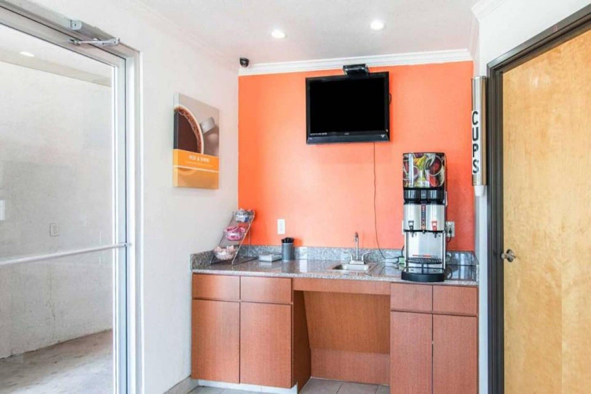 A small kitchenette with a coffee machine, a television mounted on the wall, and a door labeled 'Cups'.