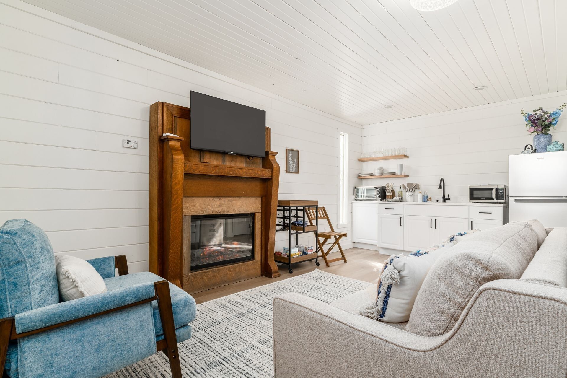Una sala de estar con chimenea, un televisor montado encima y una zona de cocina con electrodomésticos modernos.