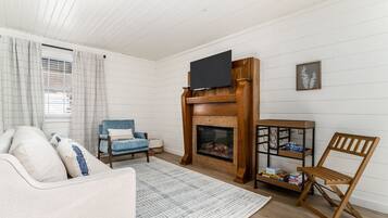 Ein Wohnzimmer mit Kamin, einem darüber montierten Fernseher, einer Couch, einem Sessel und einem Holztisch mit Büchern.