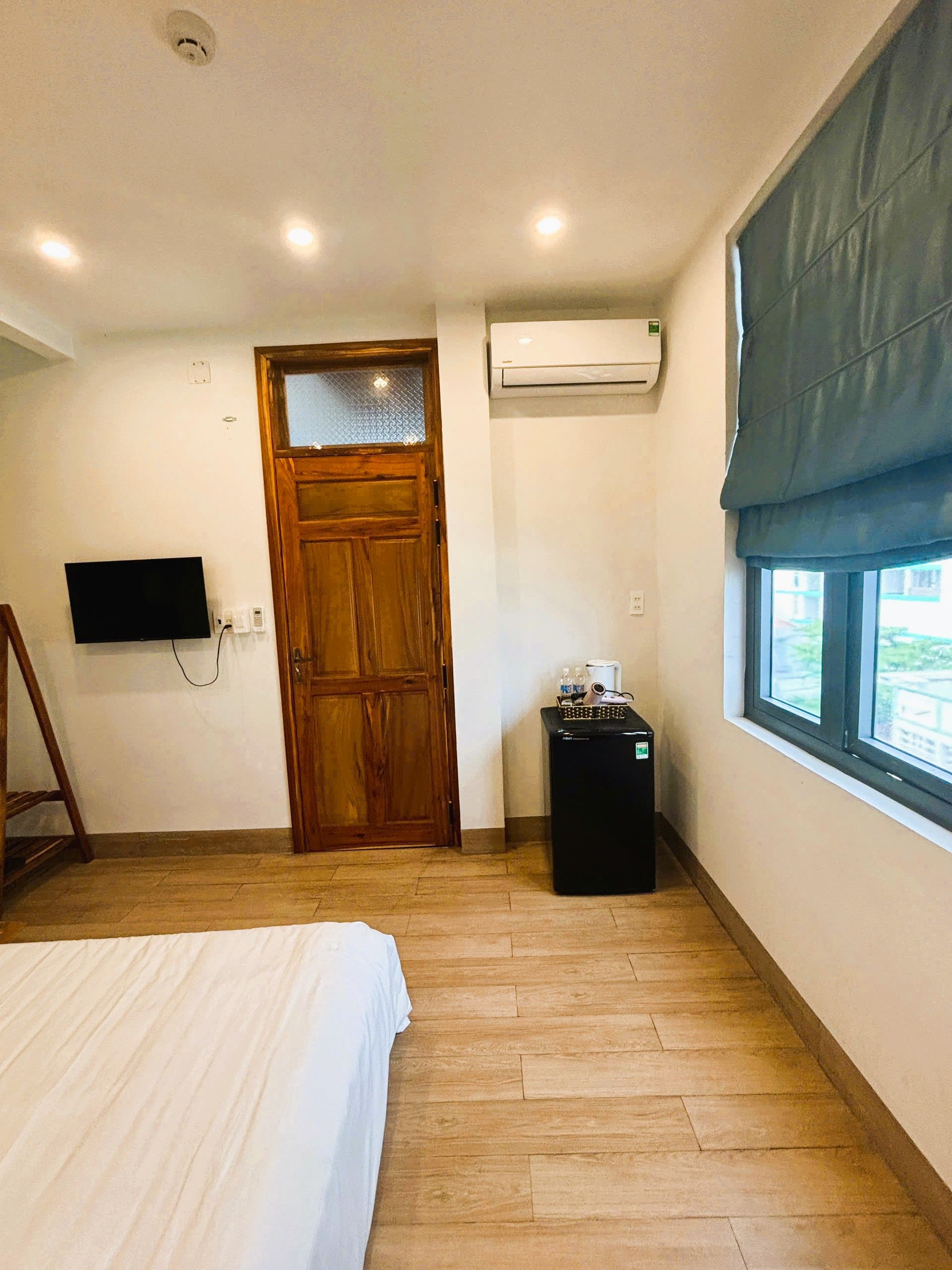 Une chambre à coucher avec un lit, une porte en bois, un appareil de climatisation, une fenêtre avec un store et une petite table sur laquelle se trouvent divers objets.