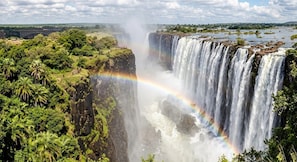 A waterfall with a visible rainbow, surrounded by lush greenery and a distant bridge. - VIEW MAPOPOMA VICTORIA FALLS HOTEL (Victoria Falls)