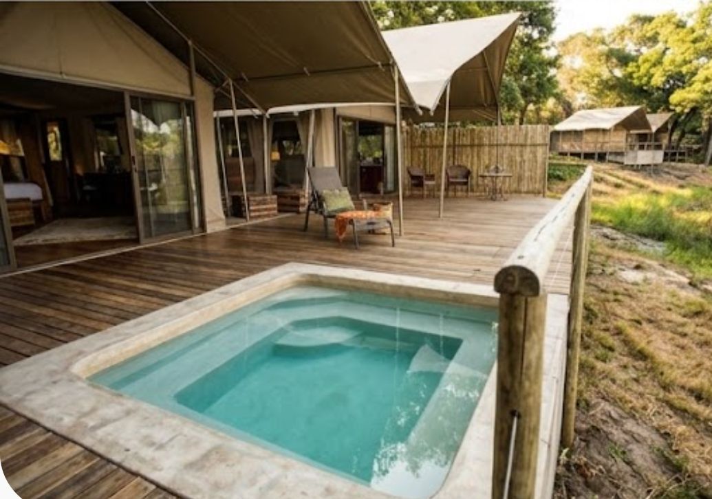 Un área de piscina con terraza de madera, zona de estar cubierta y un edificio con grandes ventanales.