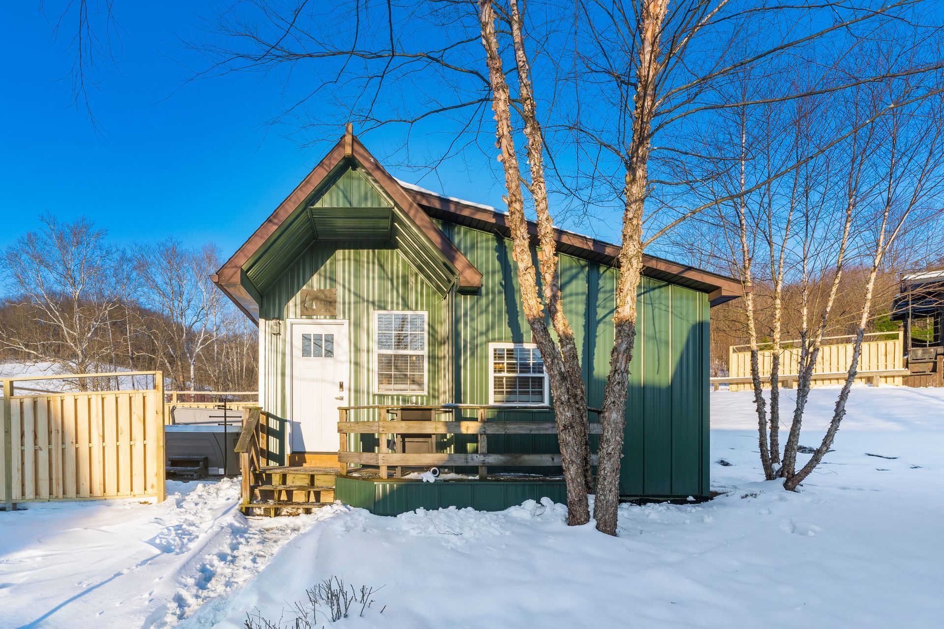 A small, green wooden cabin with a front porch and a door, surrounded by snow and trees.
