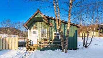 A small, green wooden cabin with a front porch and a door, surrounded by snow and trees.