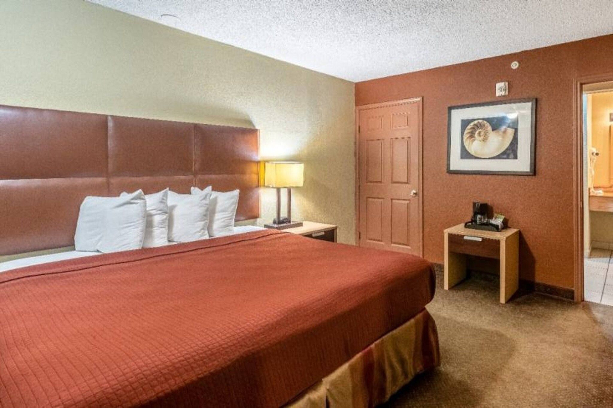 A hotel room with a large bed, a bedside table with a lamp, a framed picture on the wall, and a door leading to a bathroom.