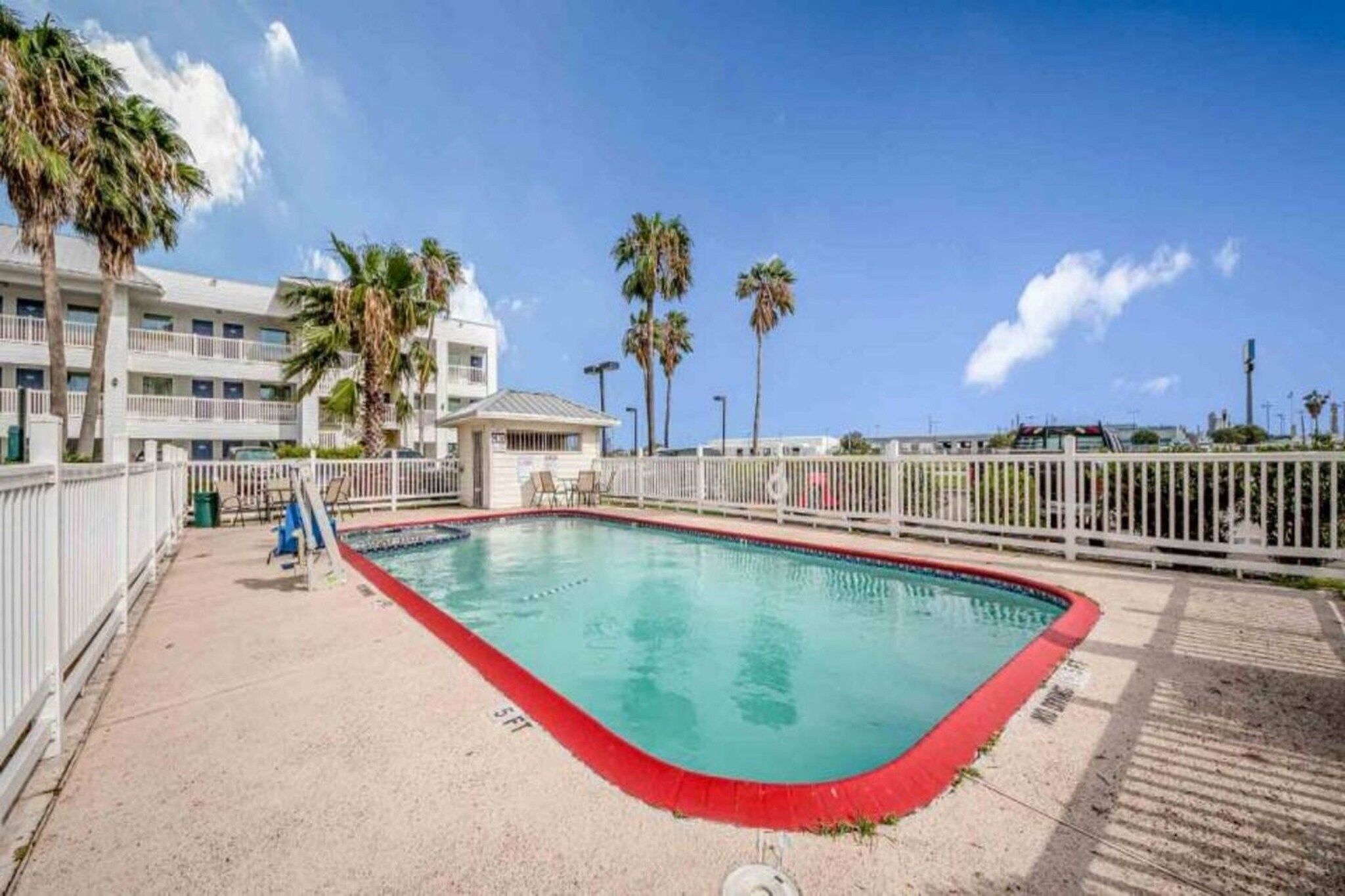 A swimming pool with a red edge, surrounded by a white fence, with palm trees and a building in the background.