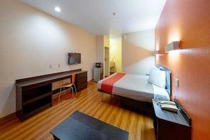 A compact hotel room with a bed, a desk, a chair, a television, and a small kitchenette.