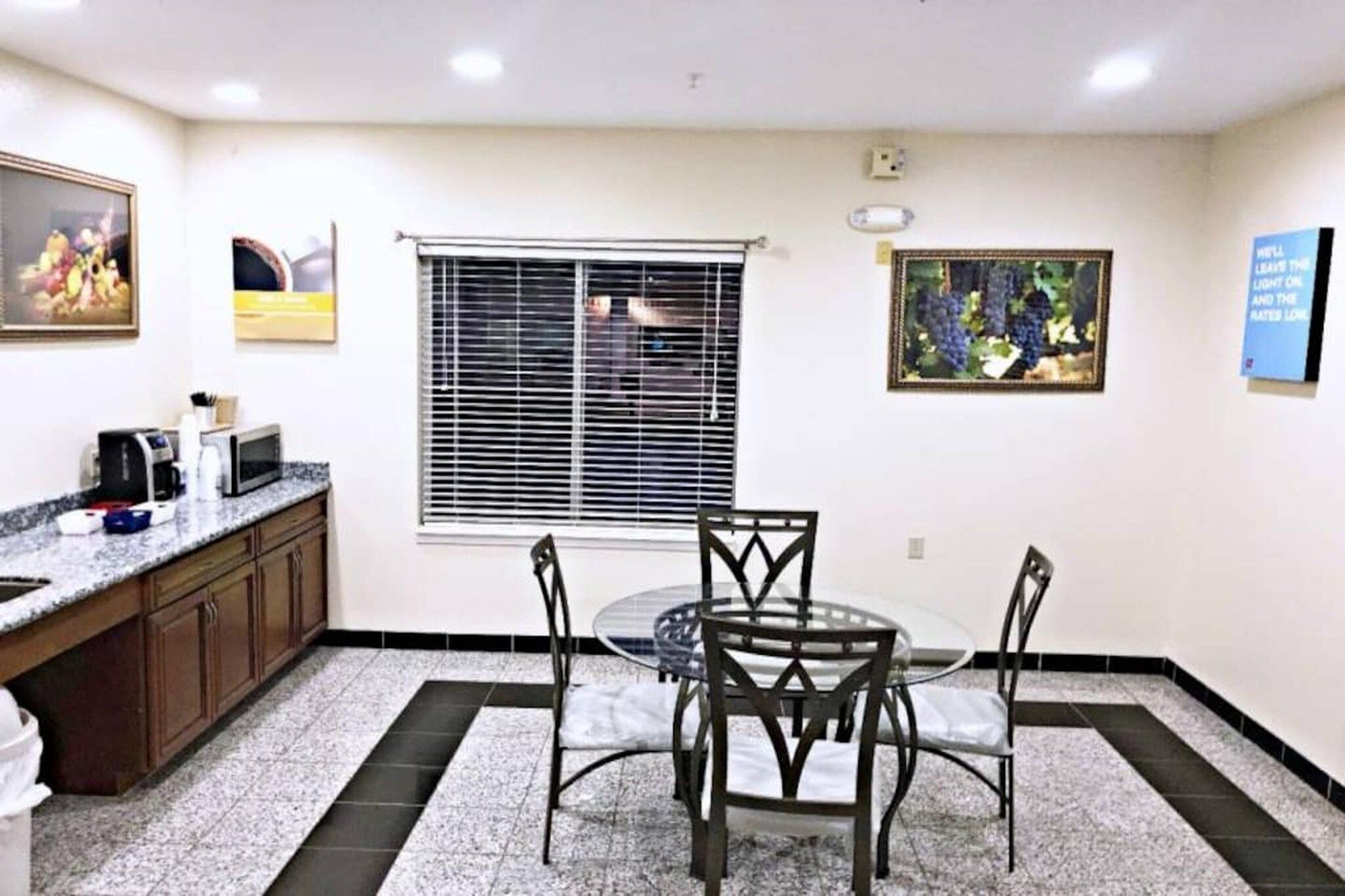 A room with a dining table, chairs, a window with blinds, a kitchen area with a microwave, and a framed picture on the wall.