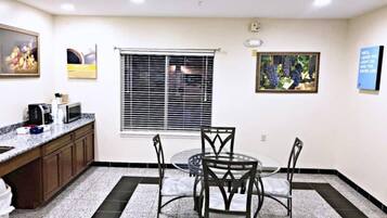 A room with a dining table, chairs, a window with blinds, a kitchen area with a microwave, and a framed picture on the wall.