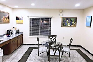 Una habitación con mesa de comedor, sillas, ventana con persianas, zona de cocina con microondas y un cuadro en la pared.