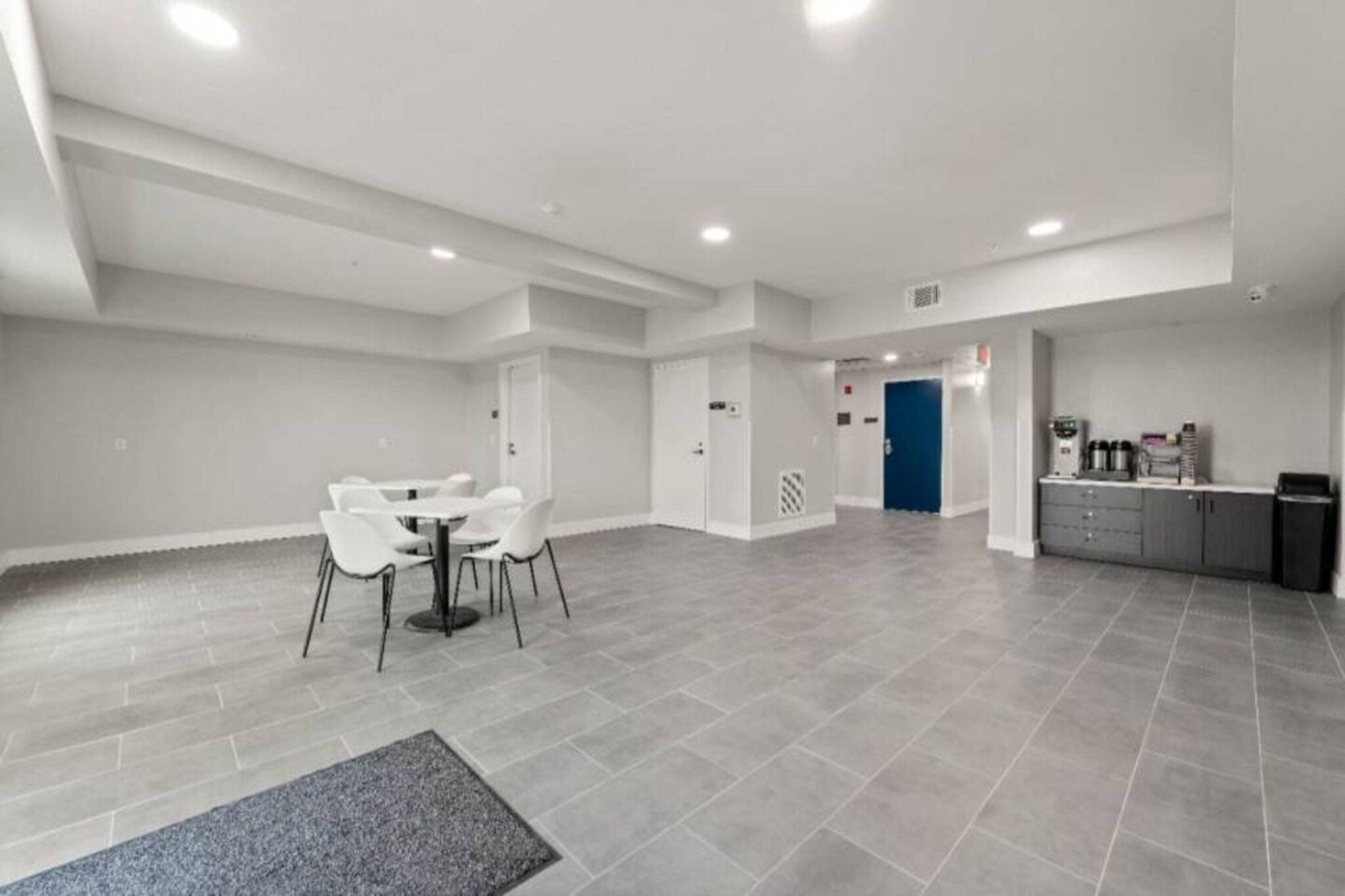 A spacious, modern hotel lobby with a dining area, a kitchenette, and a blue door leading to another room.