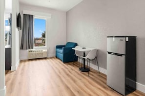 Habitació compacta amb cuina americana, zona de dinar amb taula i cadires, sofà blau i finestra gran amb cortines.