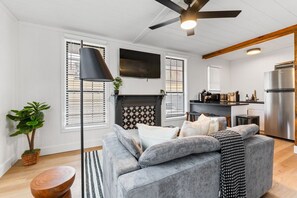 Una sala moderna con un sofá gris, una chimenea y una zona de cocina con un refrigerador de acero inoxidable.