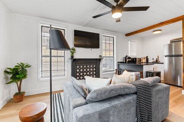 A modern living room with a grey sofa, a fireplace, and a kitchen area with a stainless steel refrigerator. - The White Lantern | Hocking, Pickleball, Hot Tub (Creola)