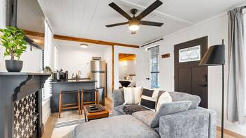 Ein modernes Wohnzimmer mit einem grauen Sofa, Holznachttischen, einem Deckenventilator und einer Küchenzeile inklusive Kühlschrank und Spüle.