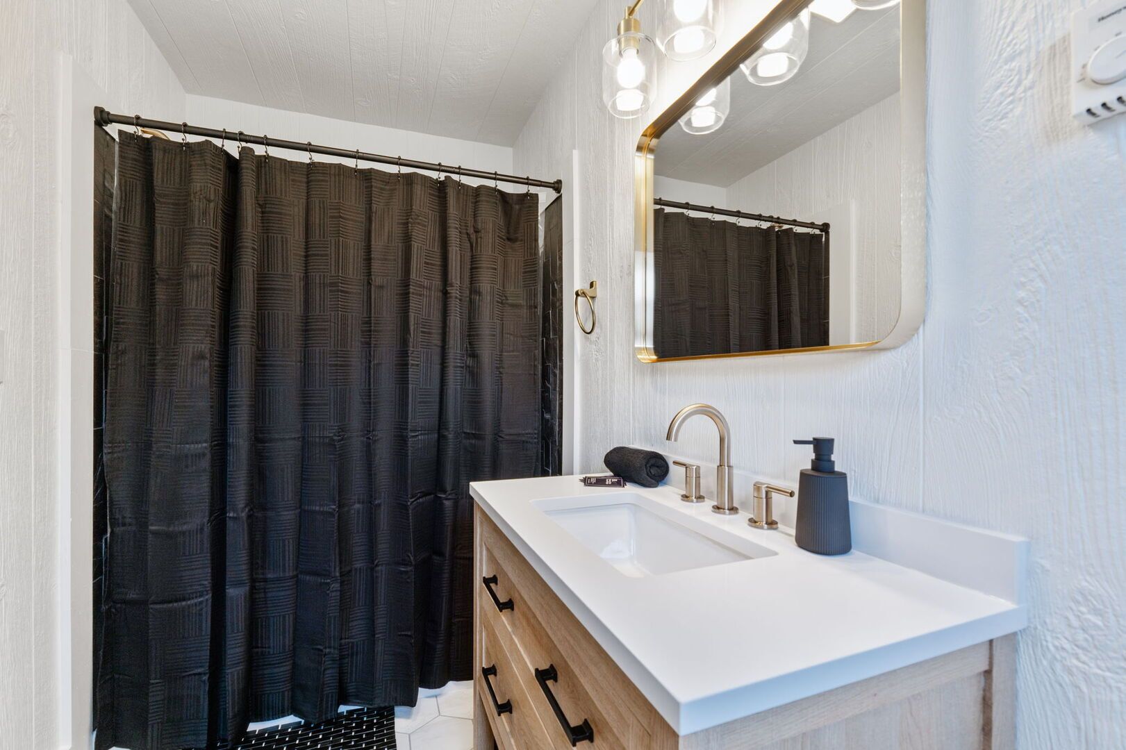 A bathroom with a white sink, a mirror with lights, and a dark shower curtain.