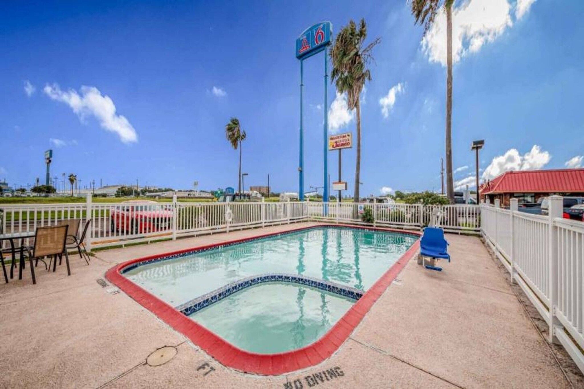 A swimming pool with a white fence, a red roofed building, and palm trees in the background.