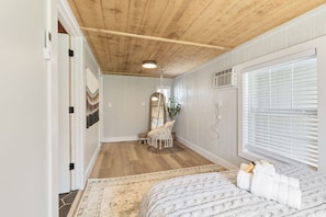 Une pièce avec un plafond en bois, un fauteuil suspendu, un lit recouvert d’une courtepointe à motifs et une fenêtre avec des stores.