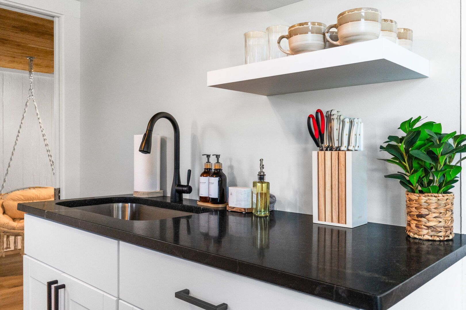 Eine moderne Küche mit schwarzem Arbeitsplatz, einer Edelstahlspüle und einer weißen Rückwand. Über der Spüle befinden sich zwei Regale, eines mit Tassen und das andere mit Küchenutensilien.