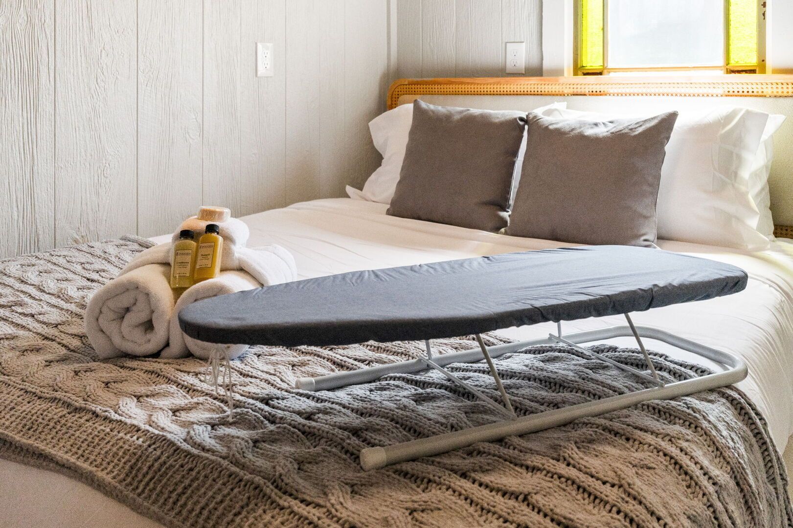 Una cama bien hecha con una tabla de planchar gris, una toalla enrollada y artículos de aseo a un lado.