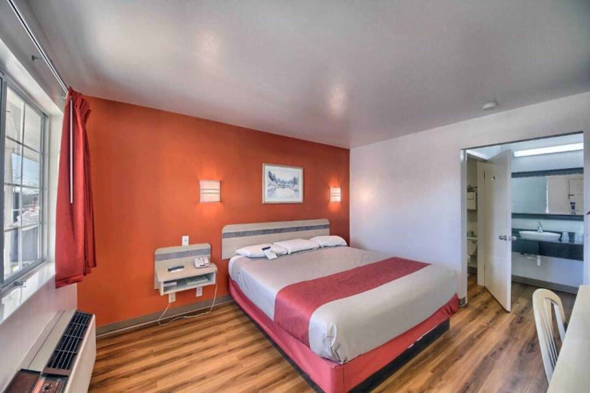 A hotel room with a bed, a bedside table, a window with curtains, a wall-mounted light, and a bathroom visible through an open door.