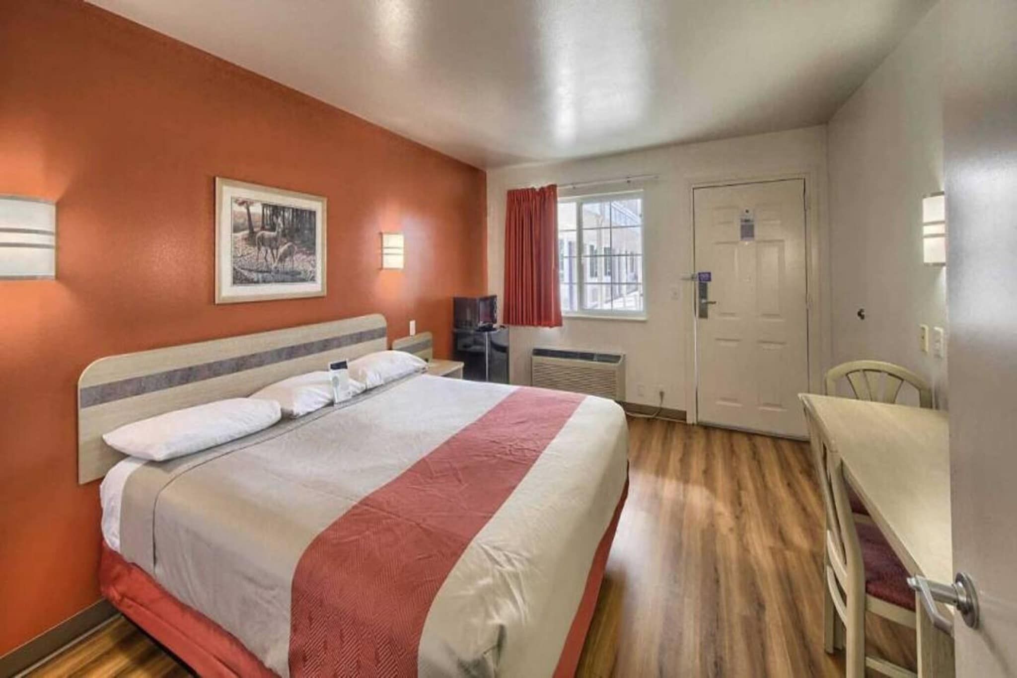 A hotel room with a bed, a desk, a chair, a window, and a door.