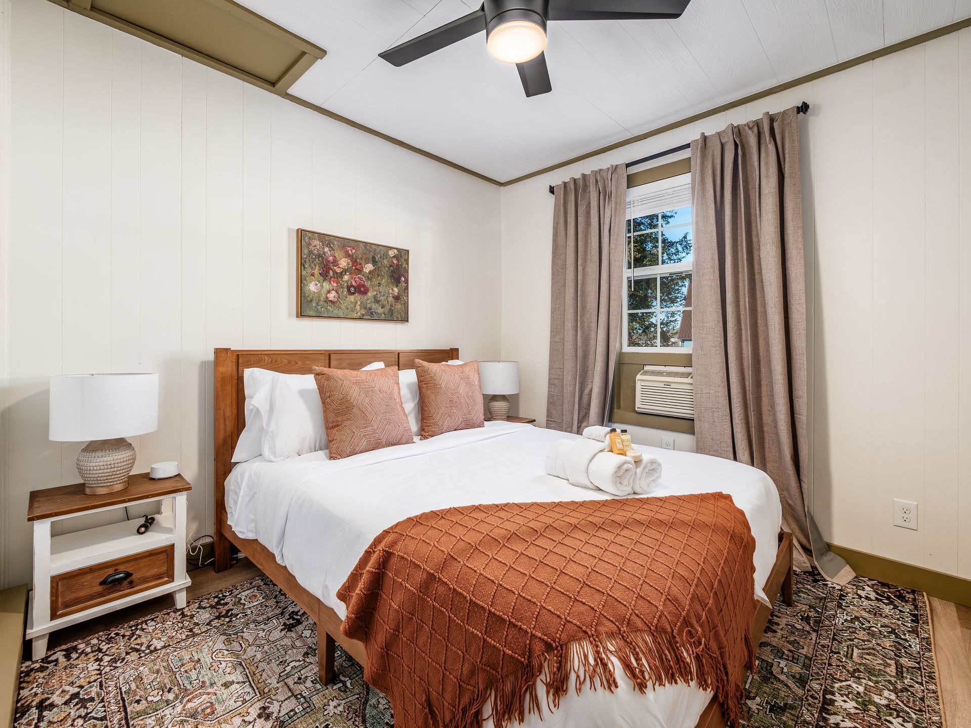 A hotel room with a bed, a nightstand, a lamp, a painting, a window with curtains, and a ceiling fan.