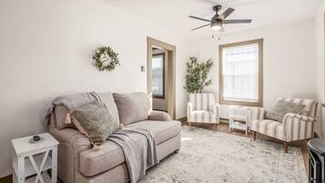 Ein Wohnzimmer mit einem beigen Sofa, gestreiften Sessel, einem kleinen Beistelltisch, einem Deckenventilator, einem dekorativen Kranz und einem großen Fenster mit Jalousien.