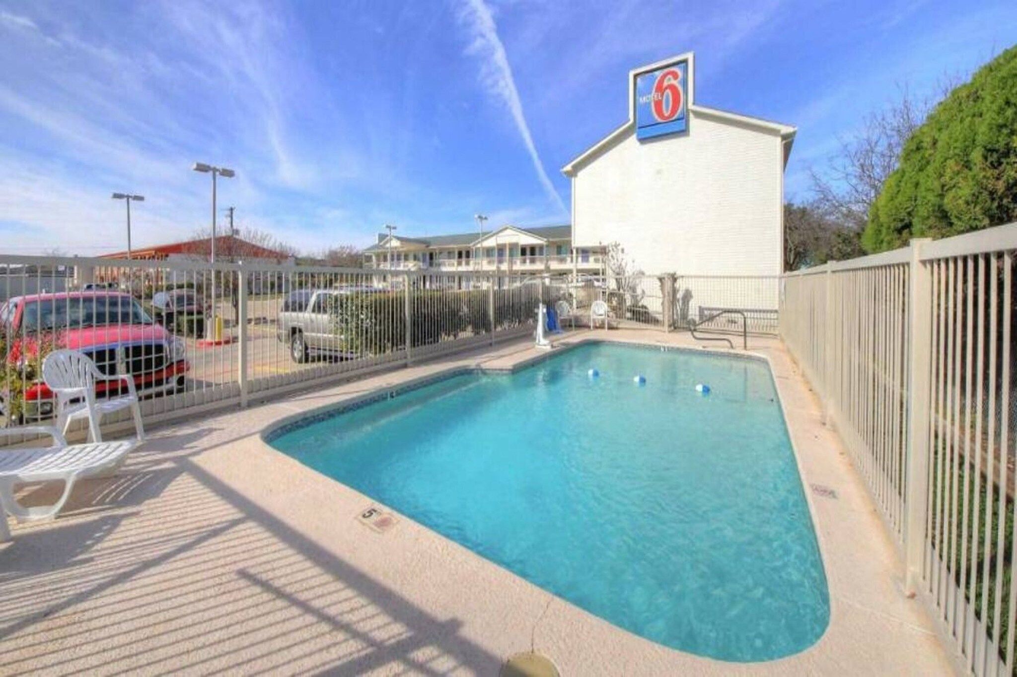 A swimming pool with a fenced area, a building with a '6' sign, and a red car parked nearby.