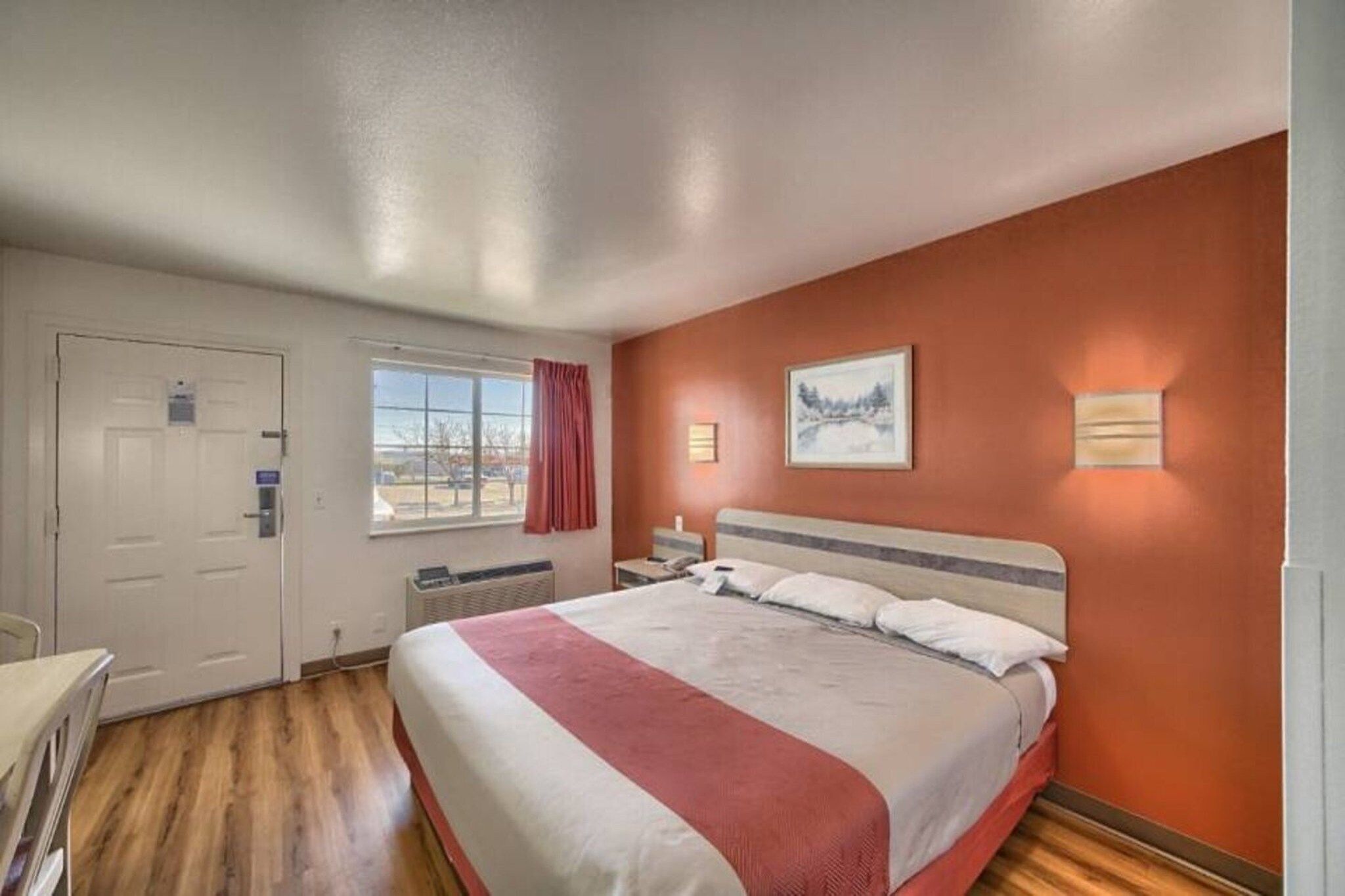 A hotel room with a bed, a desk, a chair, a window with curtains, and a painting on the wall.