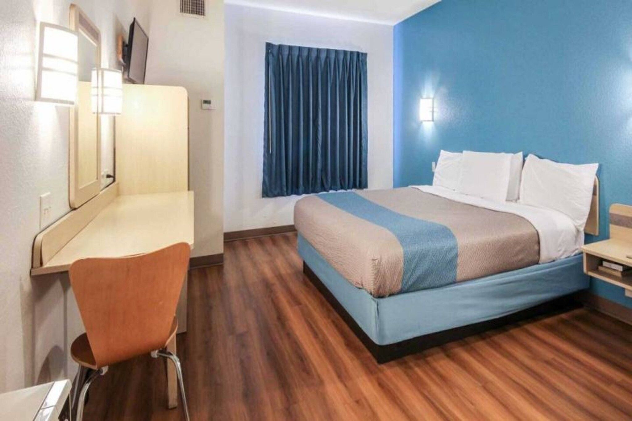 A hotel room with a bed, a desk, a chair, a window with curtains, and a wall-mounted light.