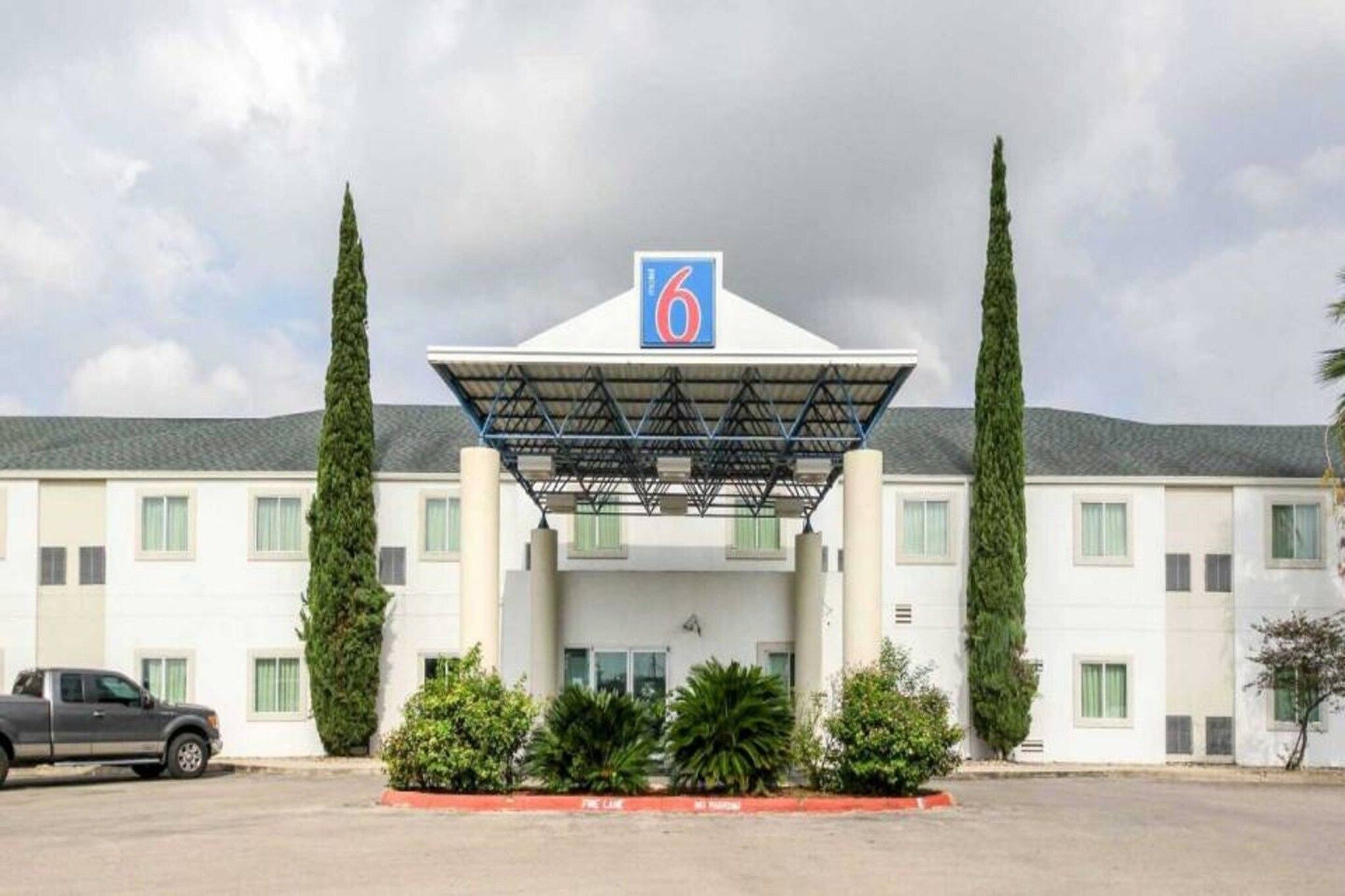 A hotel with a prominent '6' sign, a covered entrance, and symmetrical landscaping with two tall trees and several shrubs.