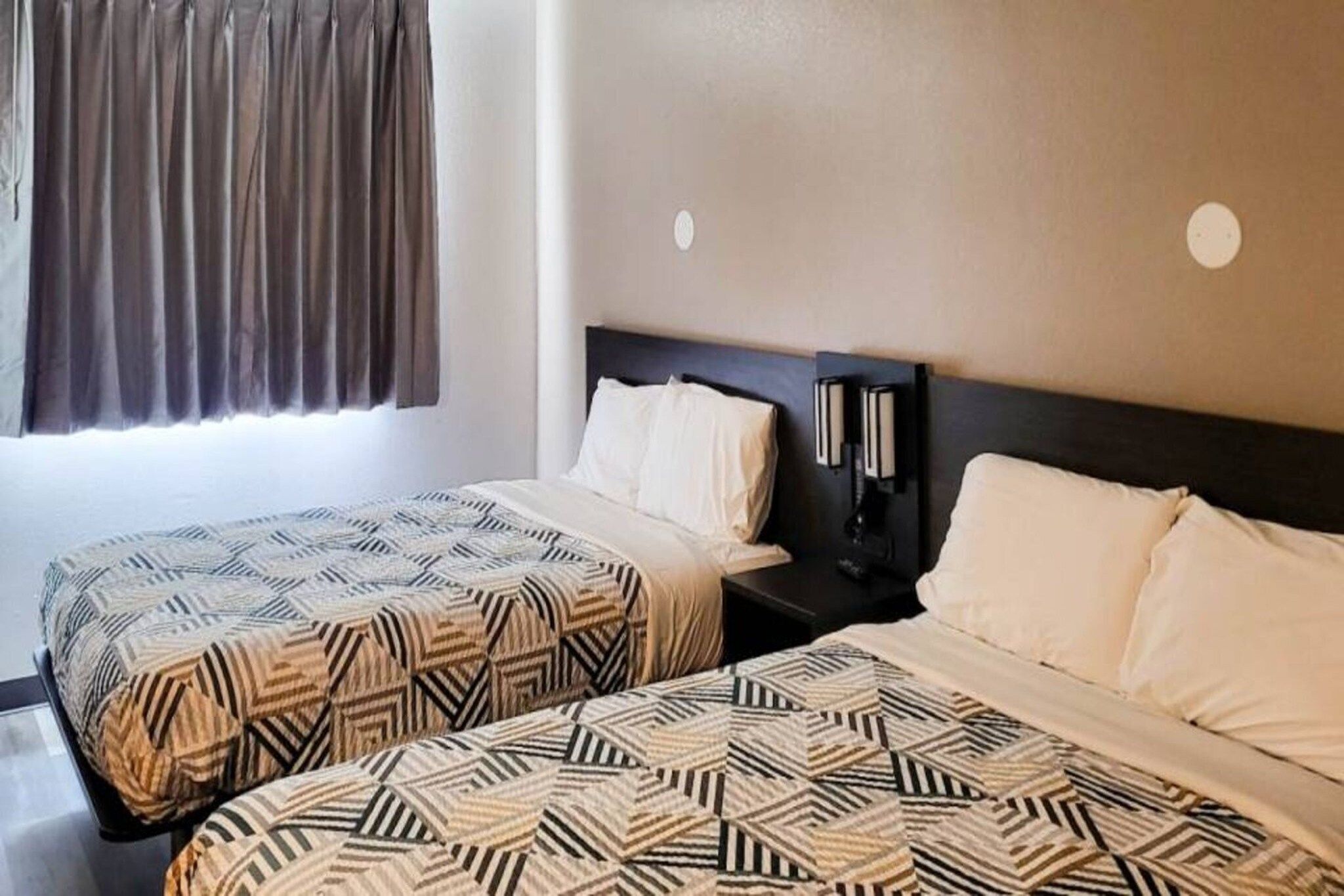 A hotel room with two beds, a nightstand, a wall-mounted phone, and a window with curtains.