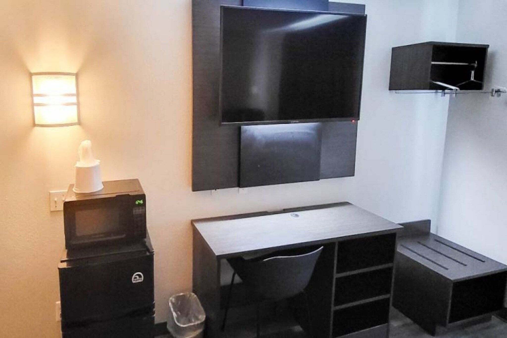 Habitación compacta con televisión de pantalla plana montada en la pared, un escritorio pequeño con una silla, un cesto de basura y un mueble de almacenamiento compacto.