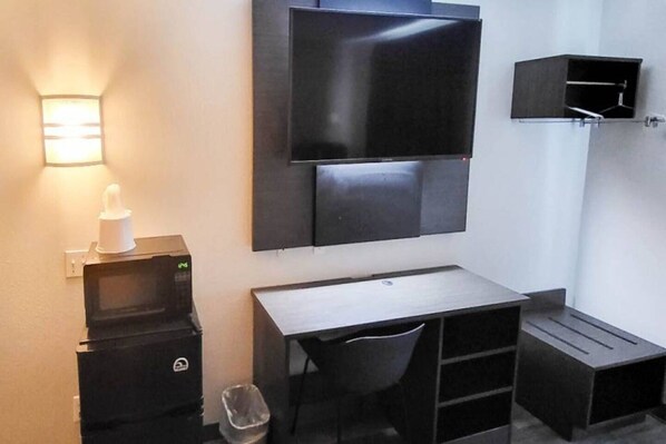 A compact room with a flat-screen TV mounted on the wall, a small desk with a chair, a trash bin, and a compact storage unit.