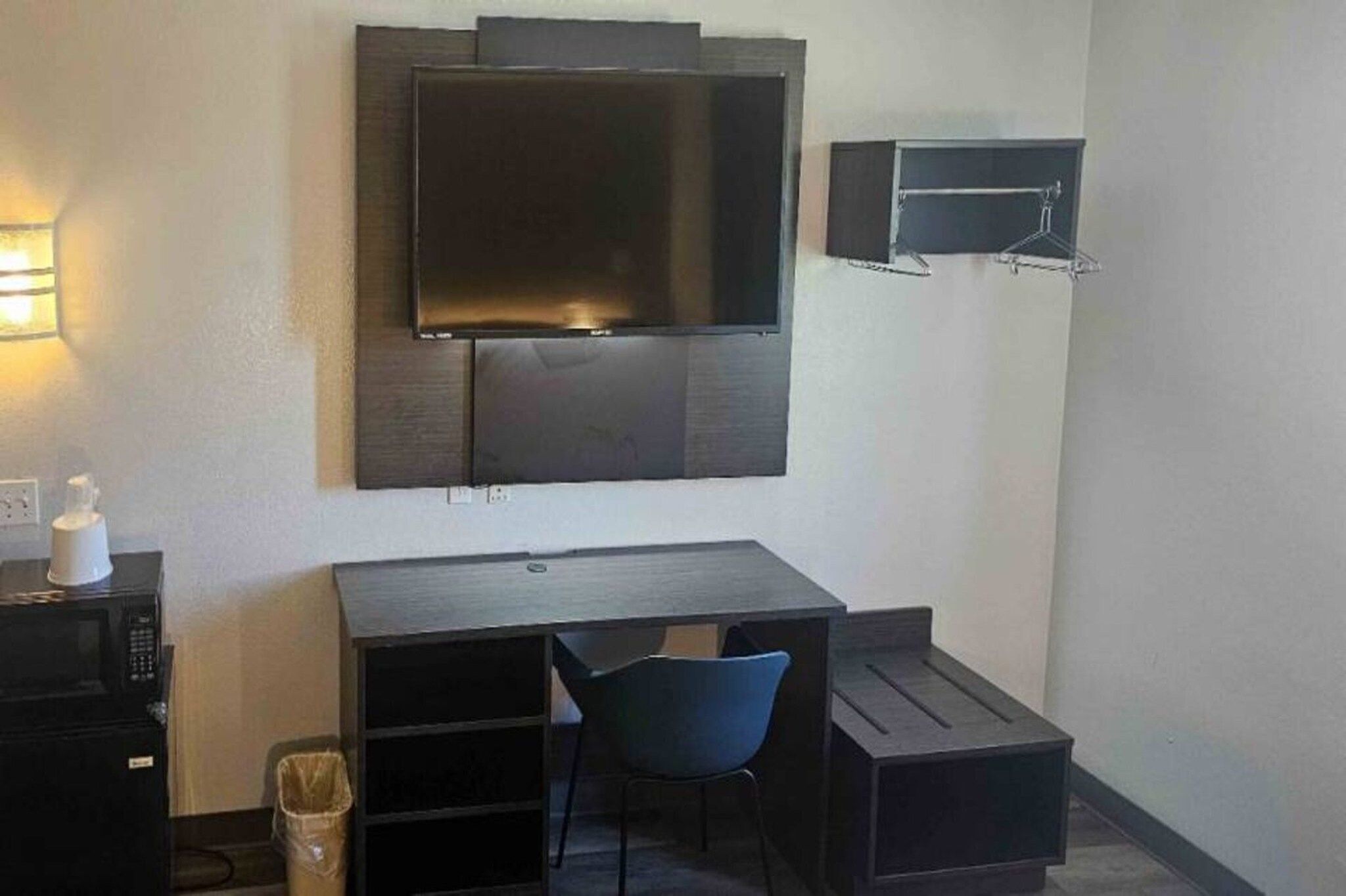A compact hotel room with a dark-colored desk, a blue chair, a television mounted on the wall, and a small shelf with a mirror.