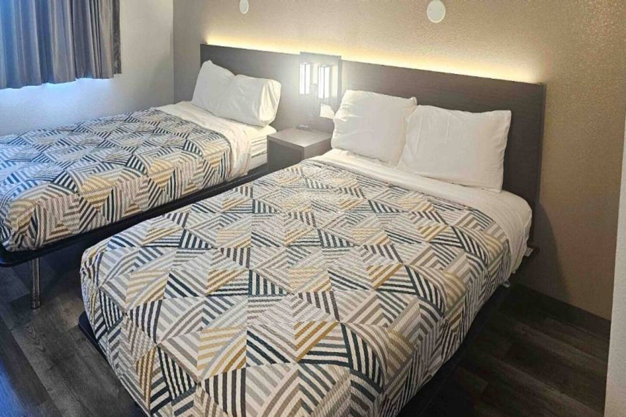 Habitación de hotel con dos camas, cada una con ropa de cama estampada, una mesita de noche y un cabecero con iluminación integrada.