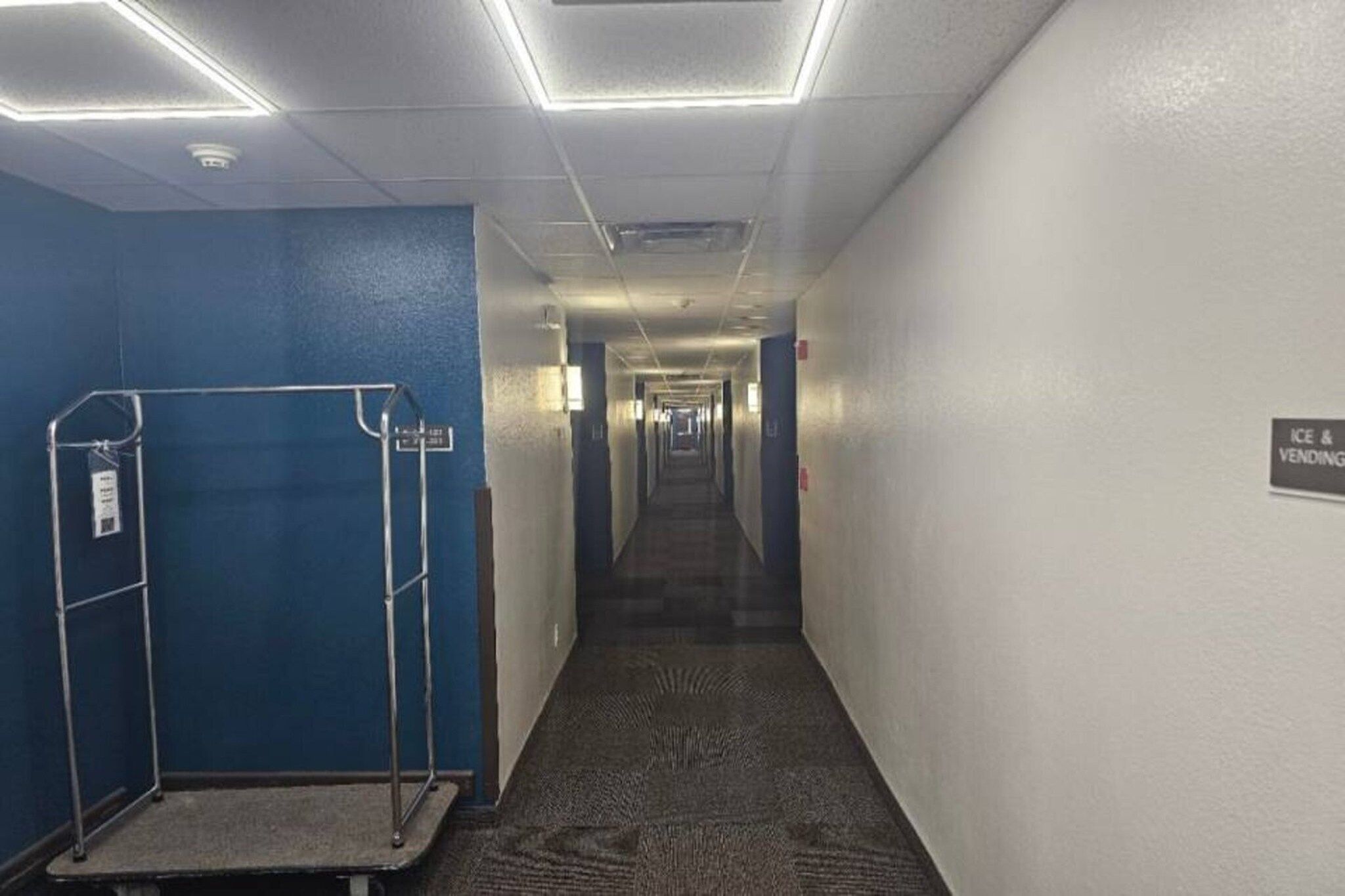 A hotel corridor with a blue wall on one side, a metal rack, and a sign indicating an ice and vending area.