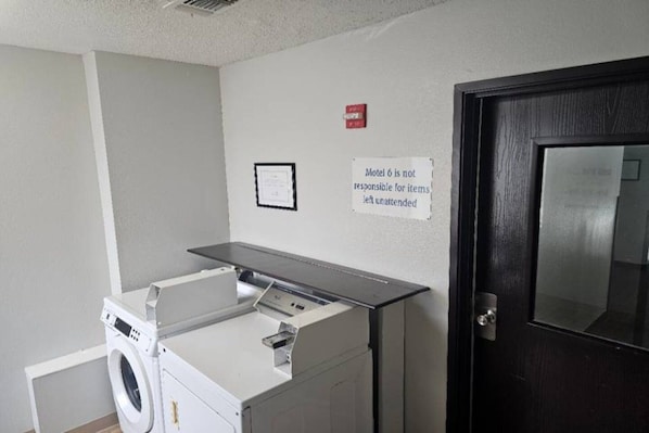 A laundry room with a washing machine, dryer, and a sign stating 'Motel 6 is not responsible for items left unattended.' - 1 Queen Bed | Non-Smoking (Sinton)
