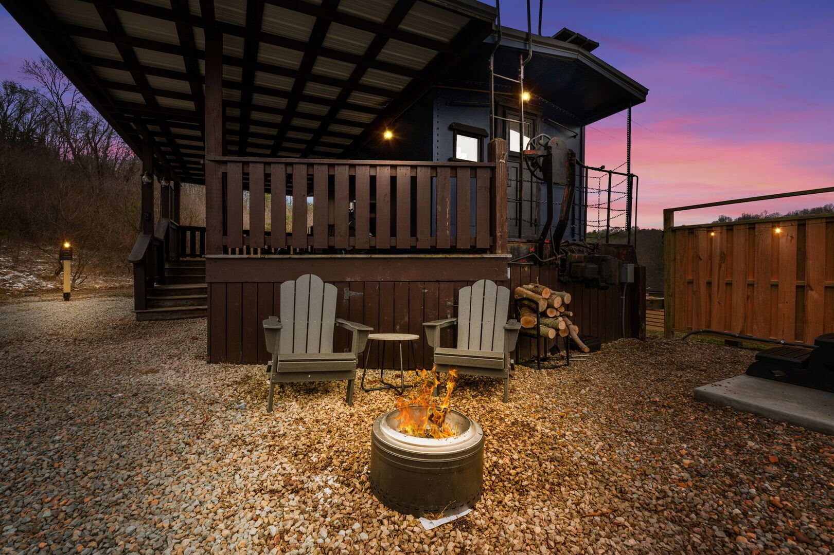 A covered outdoor seating area with Adirondack chairs and a fire pit, surrounded by a wooden deck and a gravel ground.