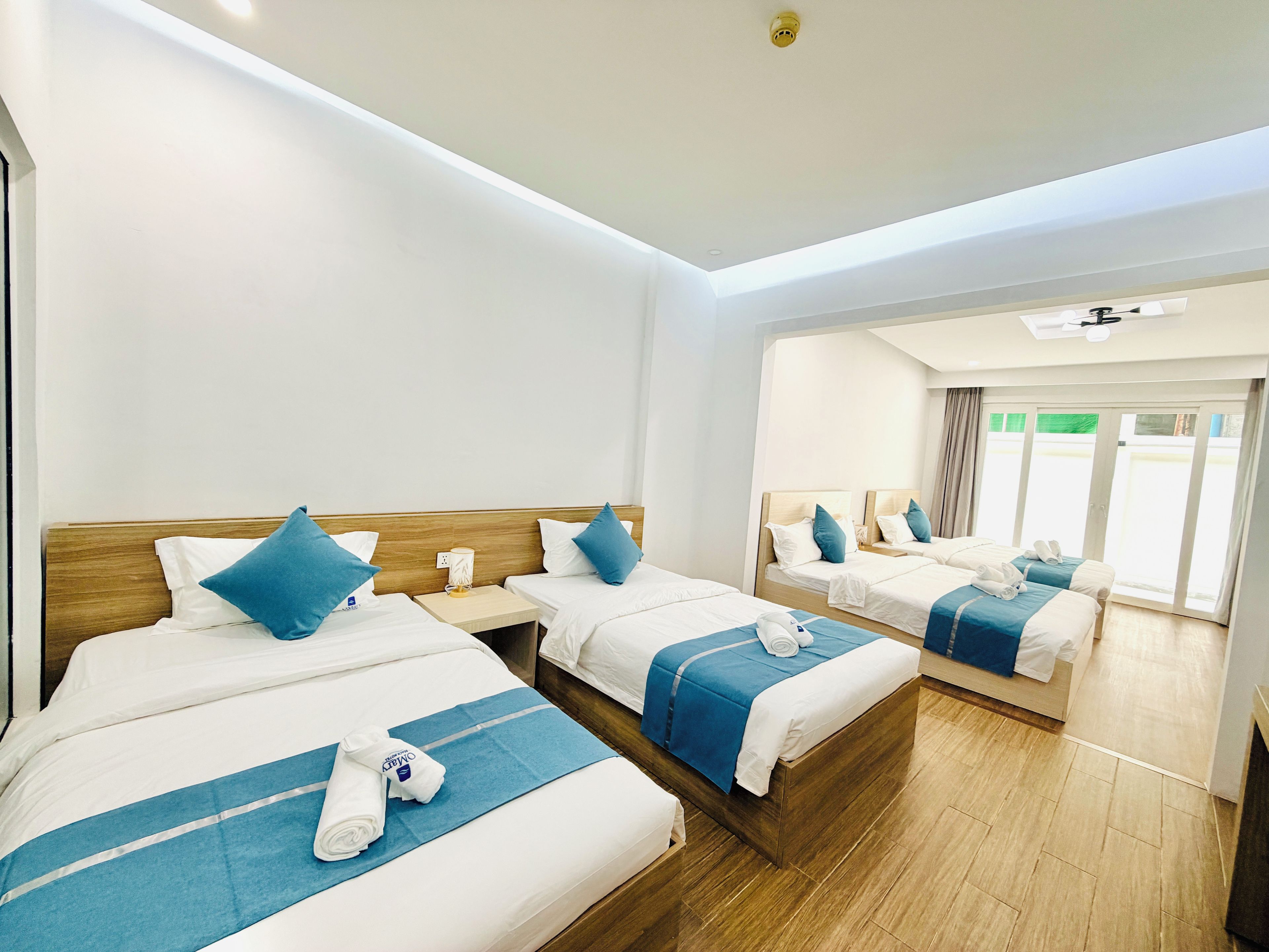 Een hotelkamer met twee bedden, elk met een houten hoofdbord, wit beddengoed en blauwe accenten. Er staan twee nachtkastjes met lampen bij, en de kamer heeft een grote schuifdeur naar een buitenruimte.