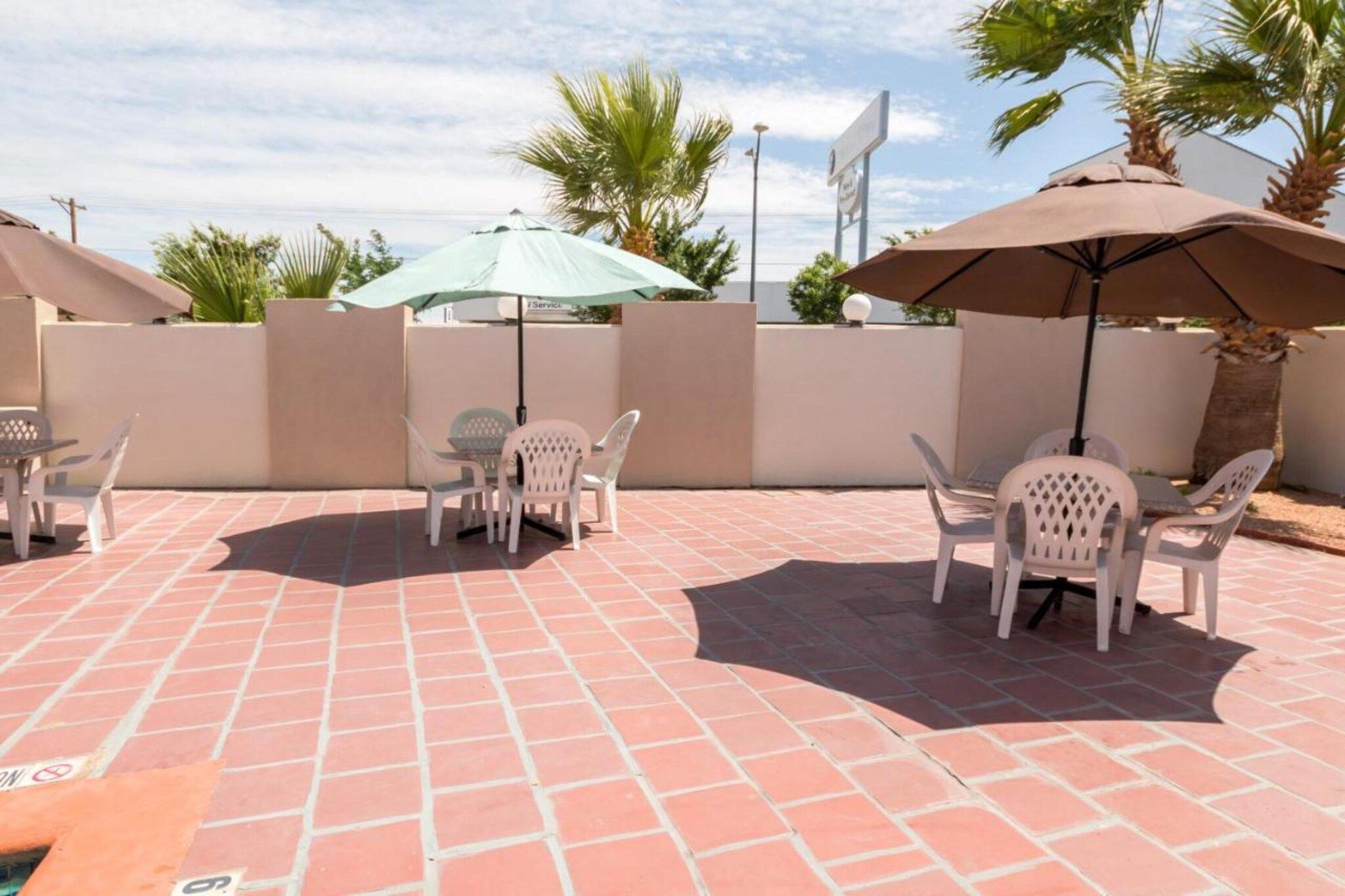 An outdoor patio with tiled flooring, white plastic chairs, and umbrellas providing shade.