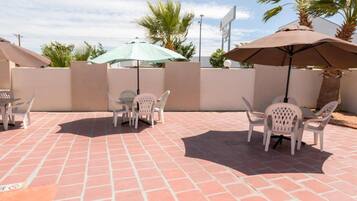 An outdoor patio with tiled flooring, white plastic chairs, and umbrellas providing shade.