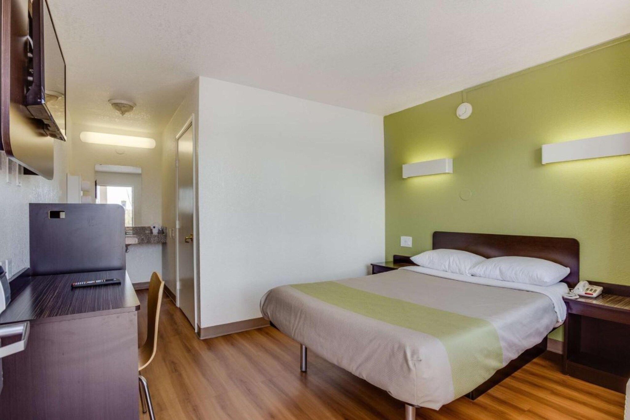 A hotel room with a bed, bedside table, lamp, and a desk with a laptop.