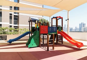 A children's play area with slides and climbing structures, set against a backdrop of a city skyline. - Waterfront Apt w/ Pool, Gym & Direct Beach Access (Sharjah)