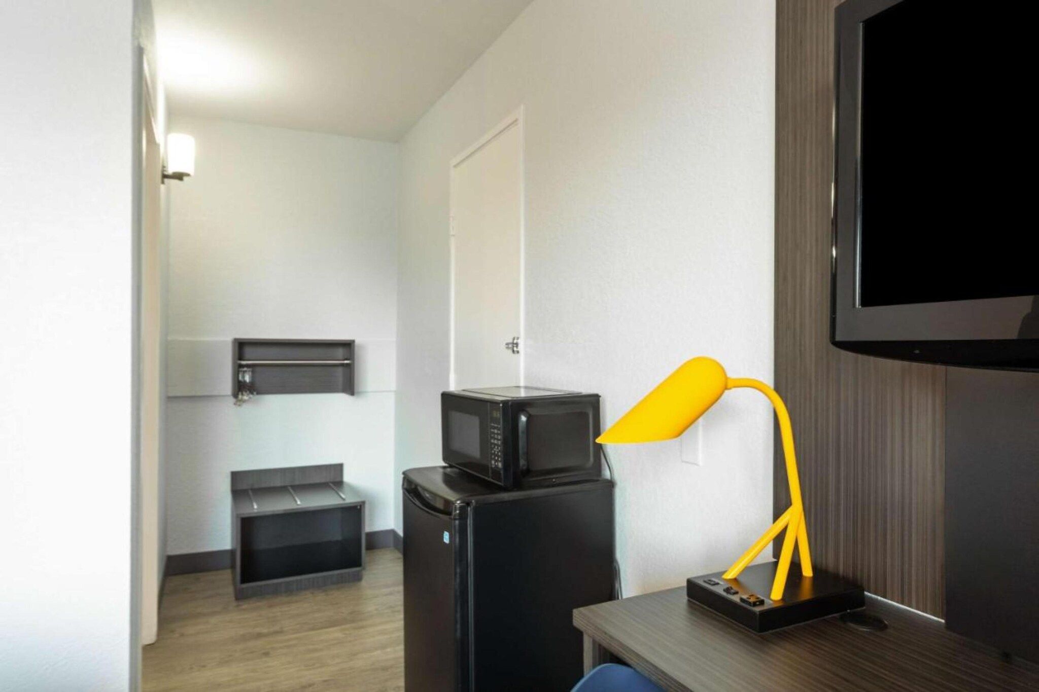 Et kompakt hotelværelse med et tekøkken, et fladskærms-tv ophængt på væggen og et lille skrivebord med lampe.