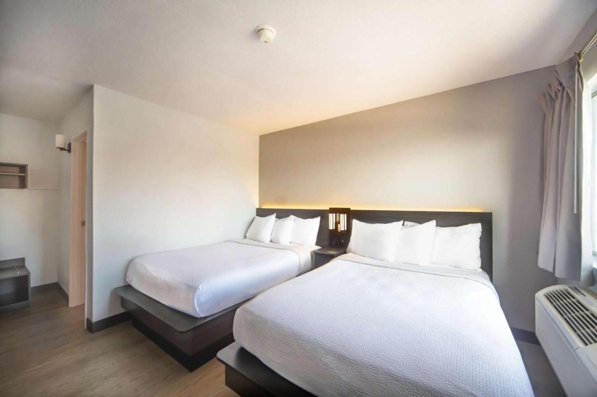 A hotel room with two beds, a bedside table with lamps, a window with curtains, and a ceiling light.