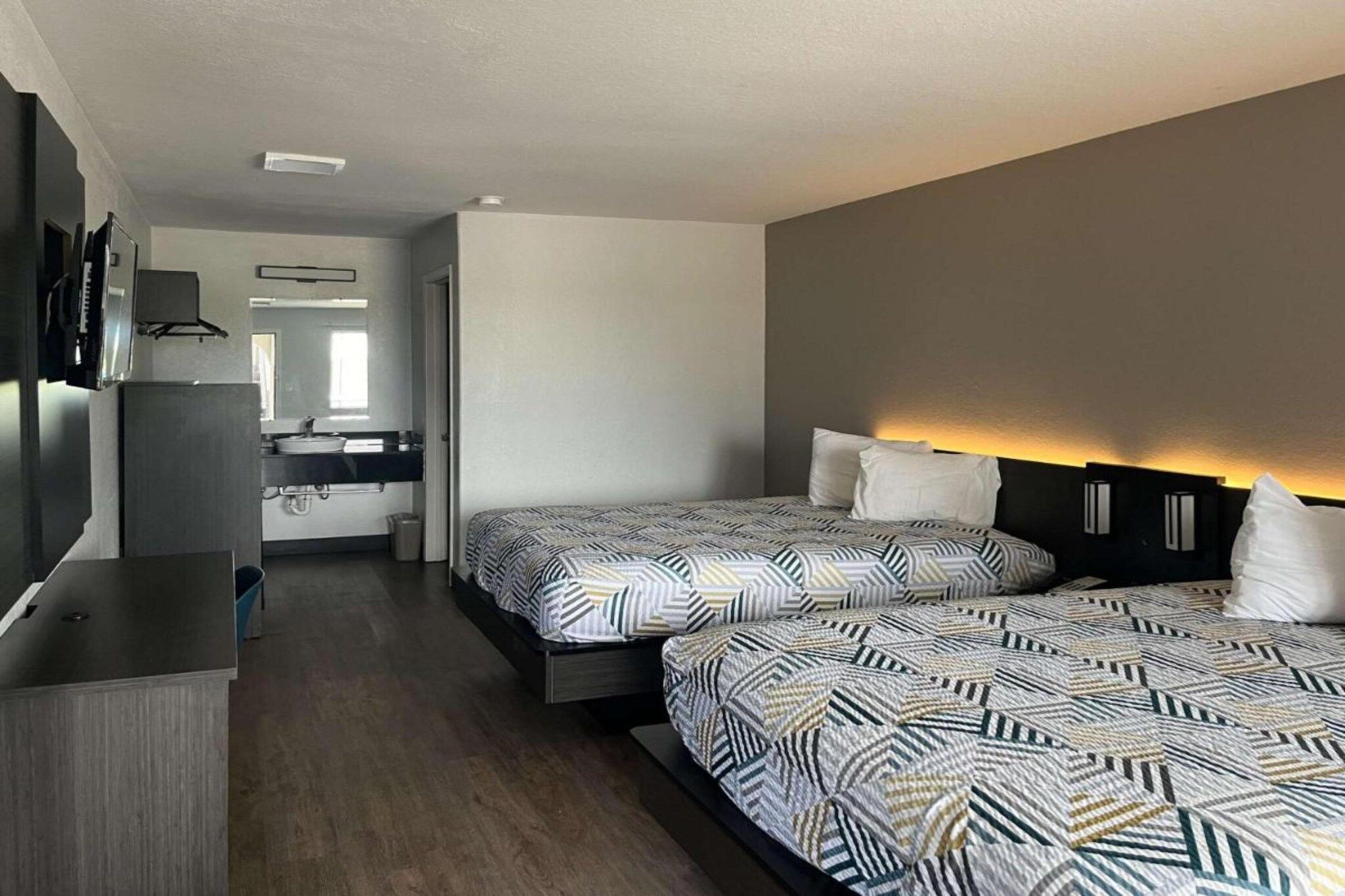A hotel room with two beds, a desk, a mirror, and a kitchenette.