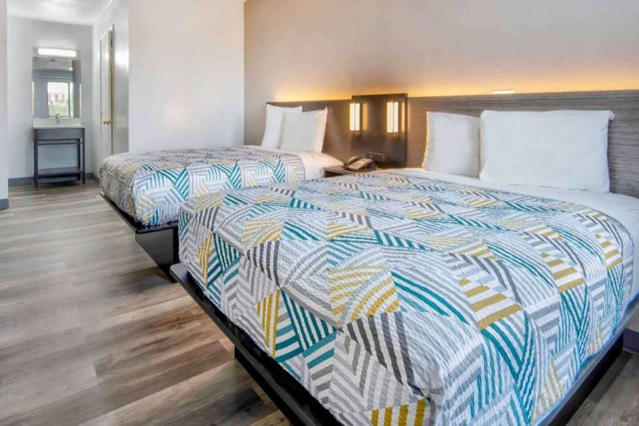 Habitación de hotel con dos camas, cada una con una colcha estampada, una mesita de noche con lámpara y vista al pasillo.